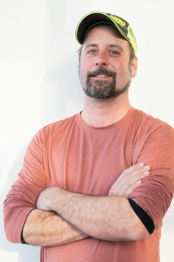 Portrait of a man with a goatee wearing a lime-green and black cap, crossed arms, and a rust-colored long-sleeve shirt against a light background.