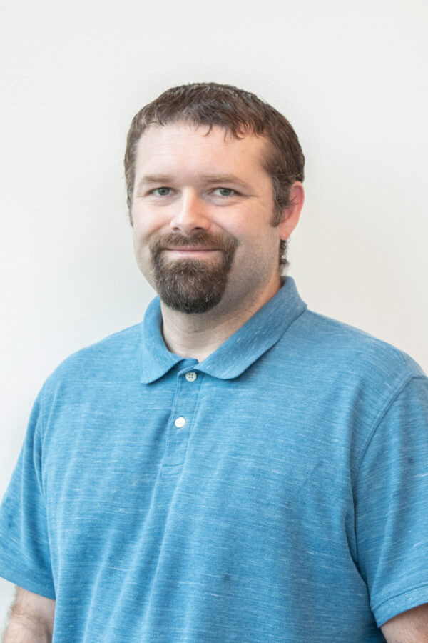 Portrait of a smiling man with short brown hair and a beard, wearing a blue polo shirt against a light background.