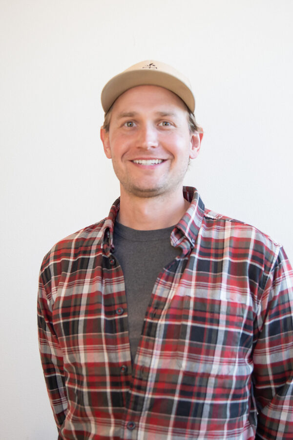 Smiling professional wears beige-brimmed baseball cap, multi-colored plaid flannel.