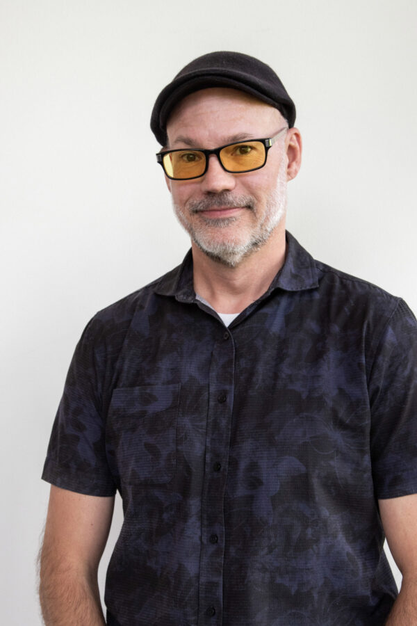 Portrait of a man wearing a black cap and amber-tinted glasses against a light background.