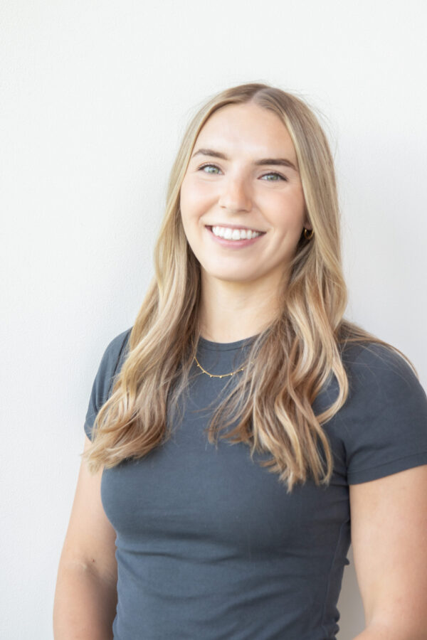Blonde woman smiles, wearing a dark gray t-shirt and subtle gold necklace. Soft light illuminates the plain white background.