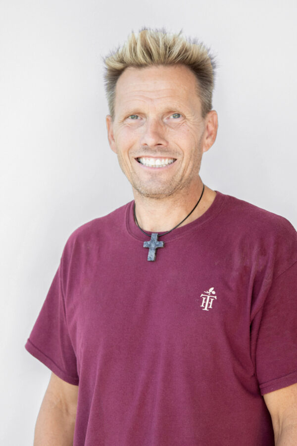 Smiling man with spiky blonde hair wearing a maroon shirt and cross necklace against a light background, in a casual portrait.