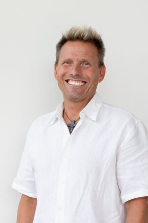 Smiling man in a white button-down shirt with a cross necklace against a light background.