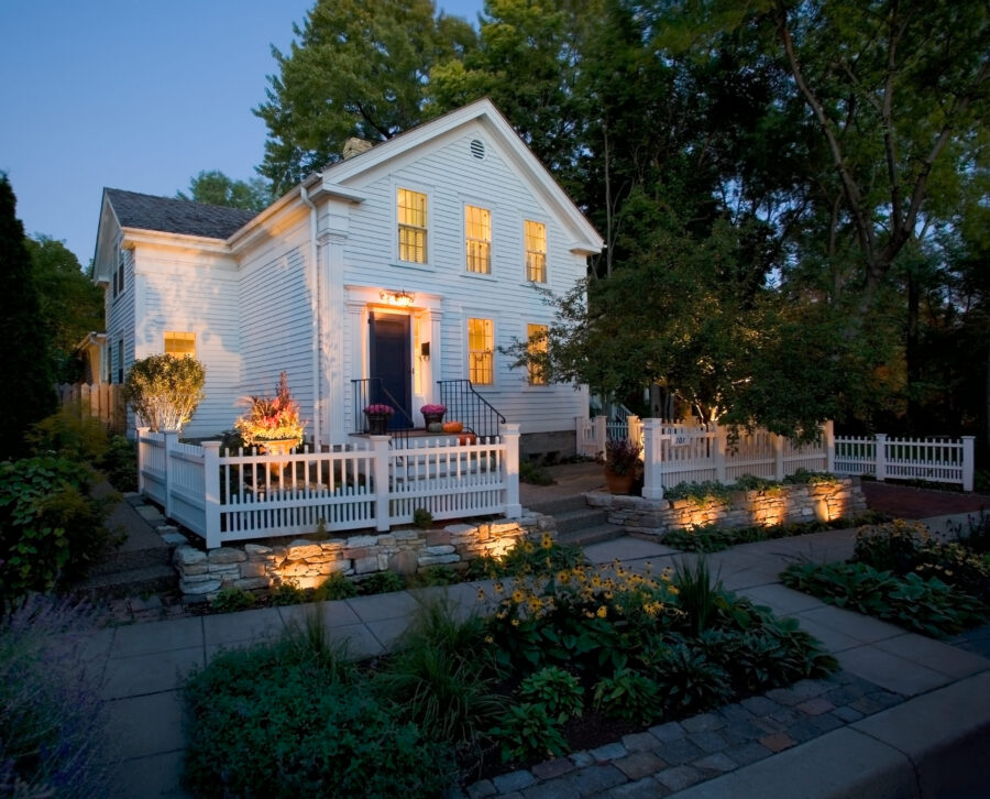 Professionally renovated home exterior features illuminated white clapboard, stone walls, and lush plantings.