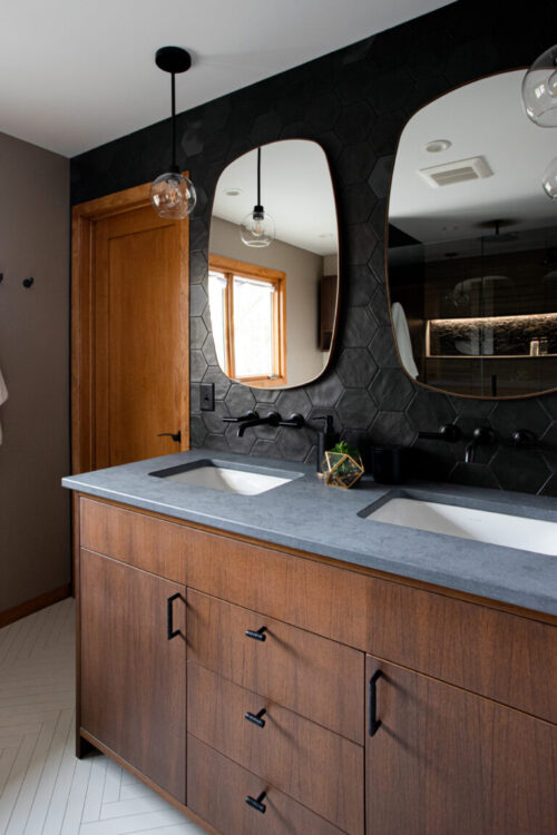 Twin Cities bathroom remodel features a dark wood double vanity, gray countertop, black hexagonal tile wall, and modern pendant lights.