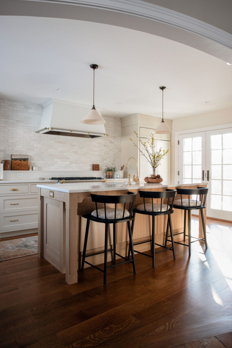 A bright Twin Cities kitchen remodel features a large light wood island and three black stools. Cream cabinets with brass hardware complement a white subway tile backsplash. Sunlight illuminates the dark hardwood floor.