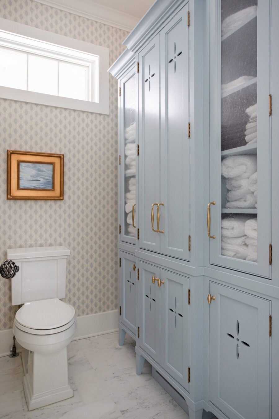 Custom light blue glass cabinetry, white toilet, patterned wallpaper, marble floor bathroom remodel.
