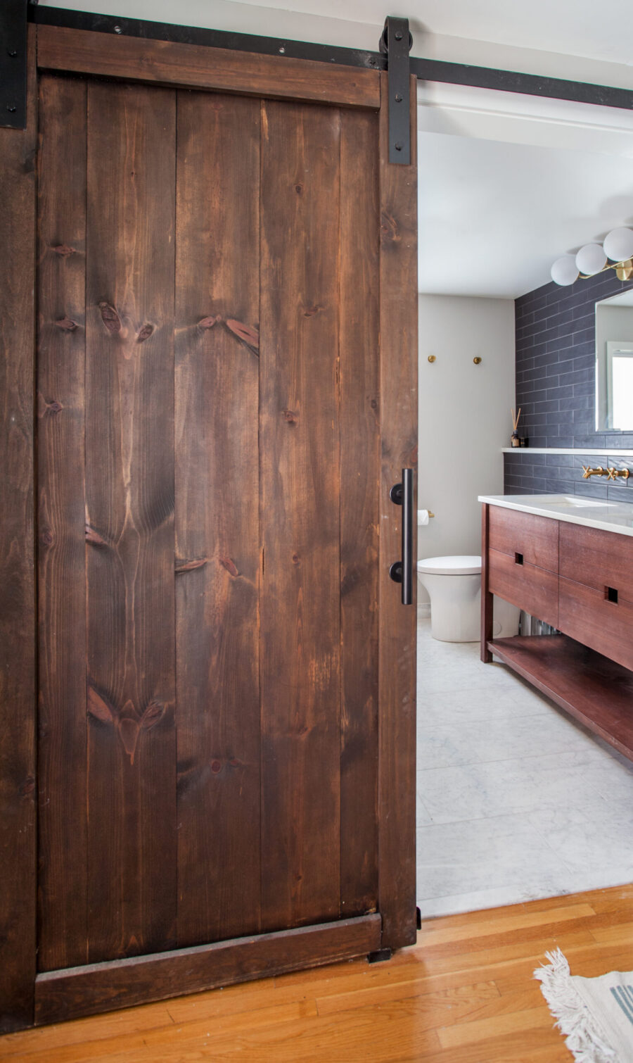 Dark wood barn door reveals a modern Twin Cities bathroom remodel: wood vanity, dark tiled wall, brass fixtures.