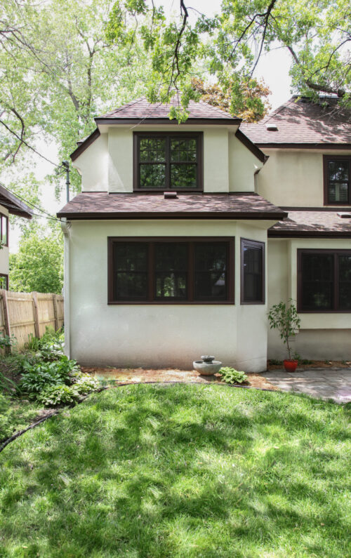 Stucco renovation features dark-trimmed windows, shingled roofs, green landscape.