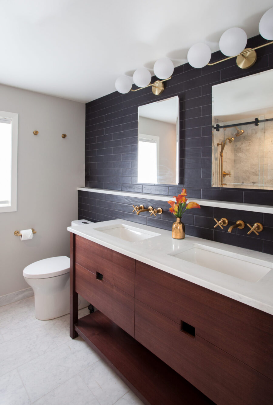 Luxurious contemporary bathroom renovation features dark tile, wood vanity, brass fittings.