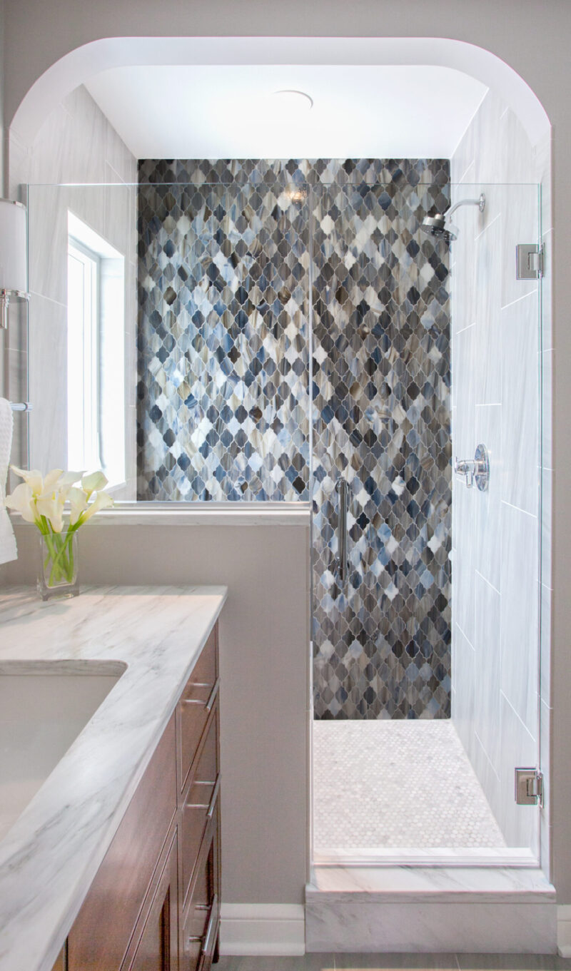 Arched frameless walk-in shower highlights patterned ogee mosaic tile, marble vanity.