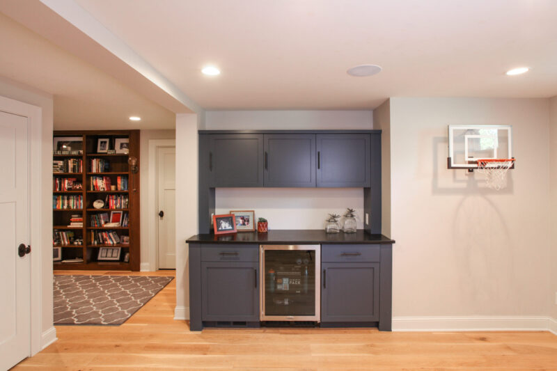 Twin Cities basement remodel features dark gray custom cabinetry, beverage fridge, light wood floors.
