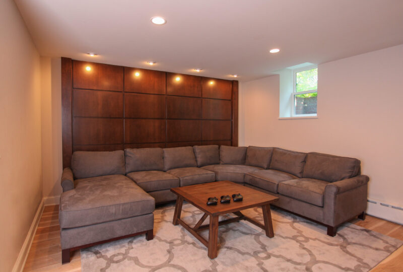 Sleek basement renovation features rich wood paneling, gray sectional, and modern lighting.