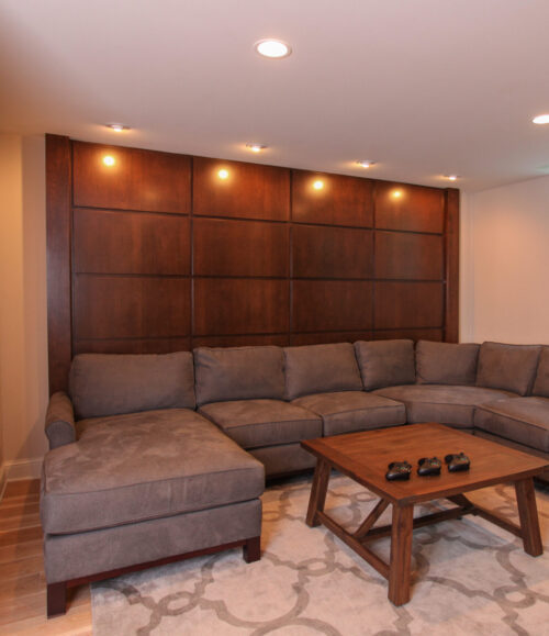 Sleek basement renovation features rich wood paneling, gray sectional, and modern lighting.