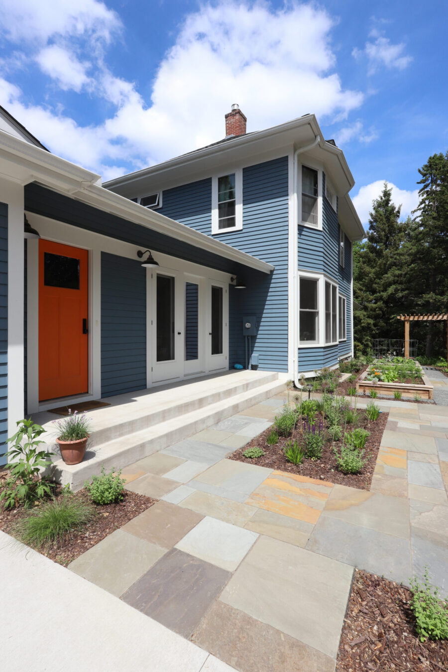 Renovated blue house exterior features an orange door, concrete steps, flagstone patio, and garden beds under a sunny sky.