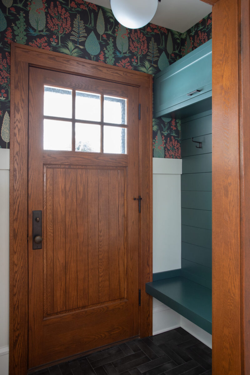 Entryway remodel. Craftsman wood door, dark patterned wallpaper, teal built-in, dark herringbone floor.