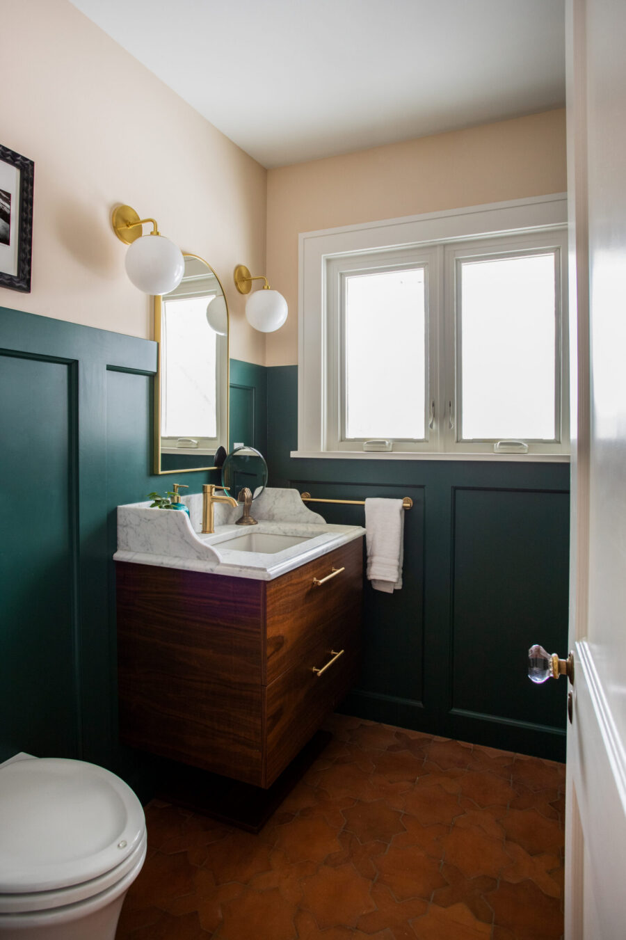 Twin Cities bathroom remodel: dark green wainscoting, floating wood vanity with marble top, brass fixtures, terracotta floor.