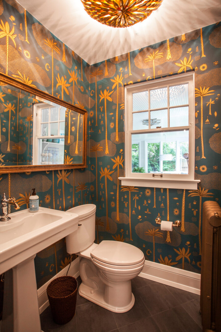 Twin Cities bathroom renovation features gold palm wallpaper, white pedestal sink, and ornate ceiling light.