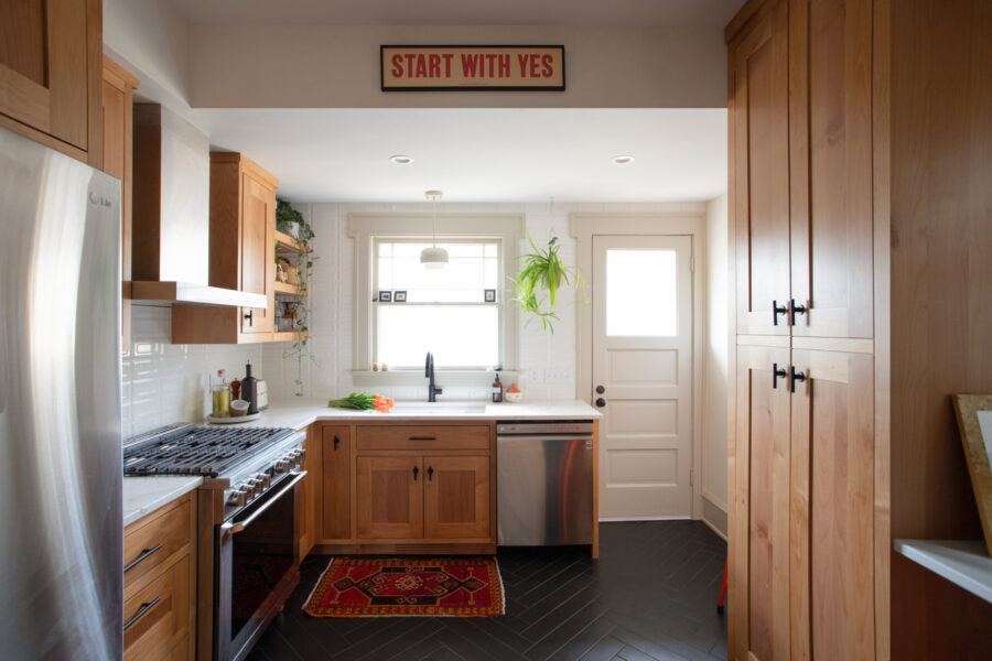 Twin Cities kitchen remodel displays light wood cabinetry, white subway tile, dark herringbone floor, and a 'START WITH YES' sign.
