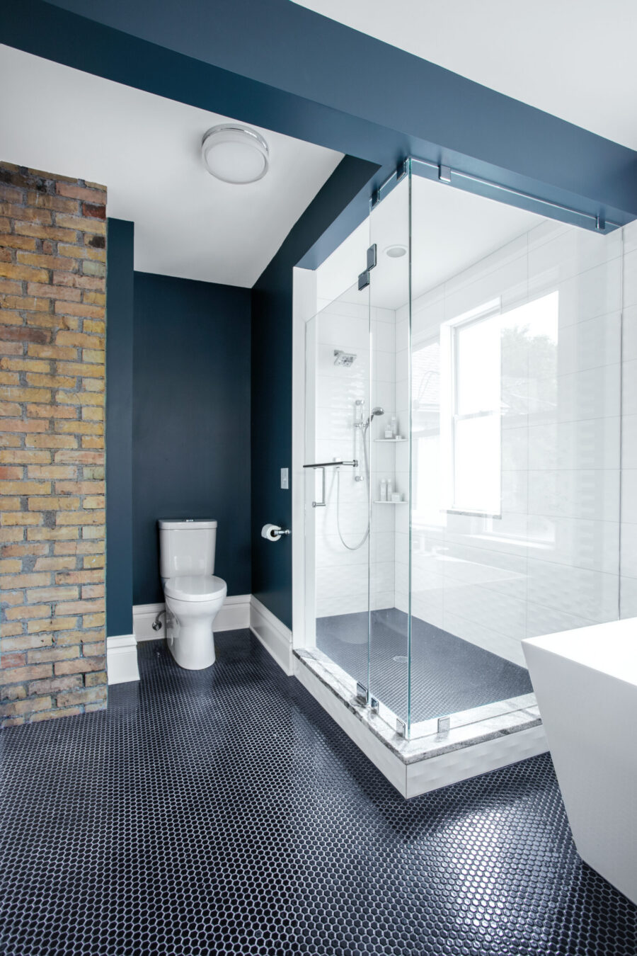 Renovated bathroom features exposed brick, dark blue walls, glass shower, and hexagonal floor tile.