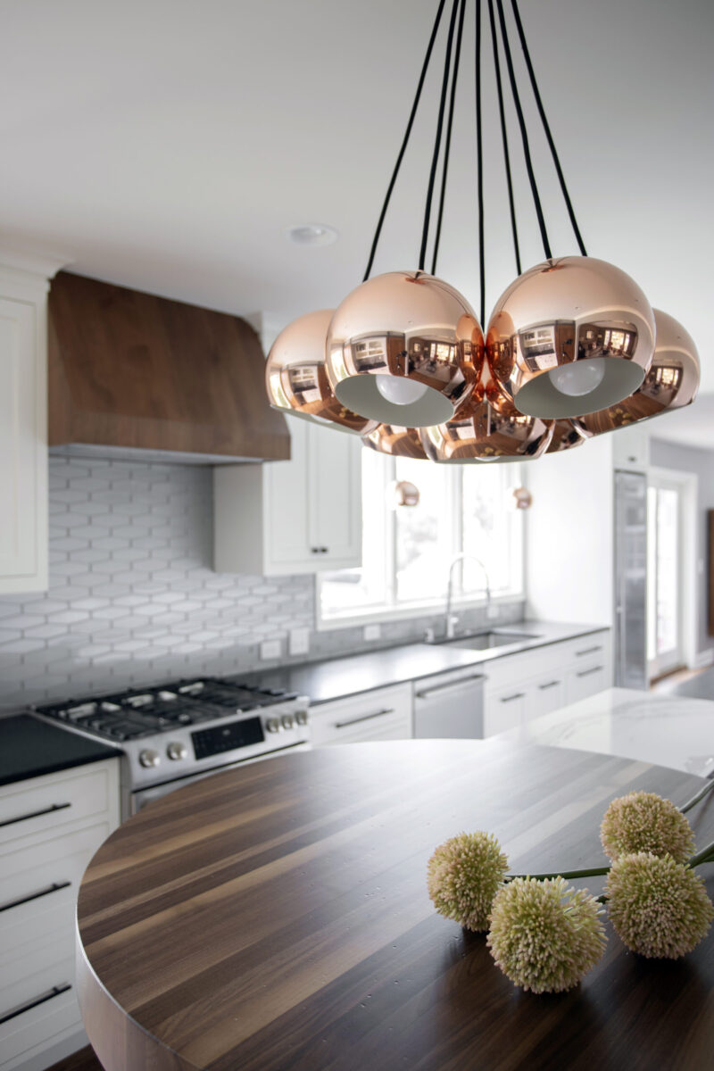 Spacious, contemporary kitchen remodel reveals white cabinetry, wood hood, and reflective copper pendants.