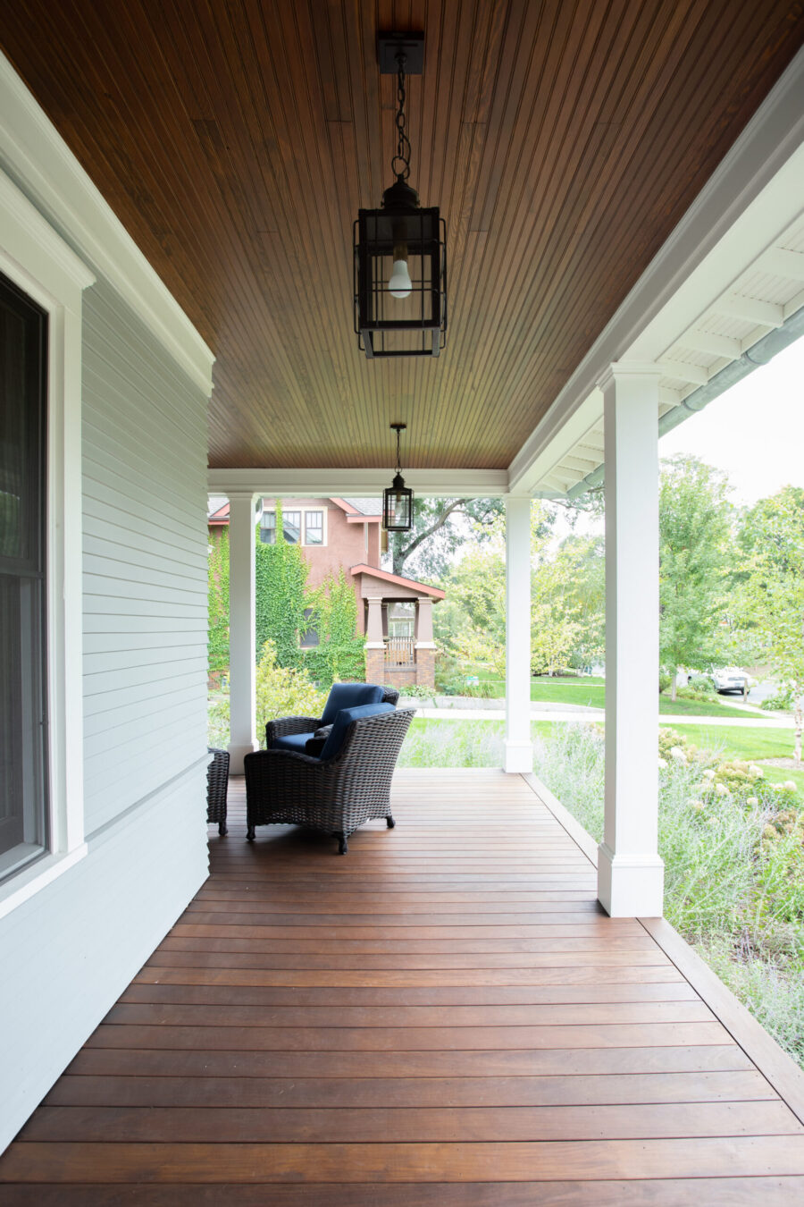 Dark-stained-wood-floored porch features beadboard-paneled ceiling, two black-framed lanterns, wicker seating.