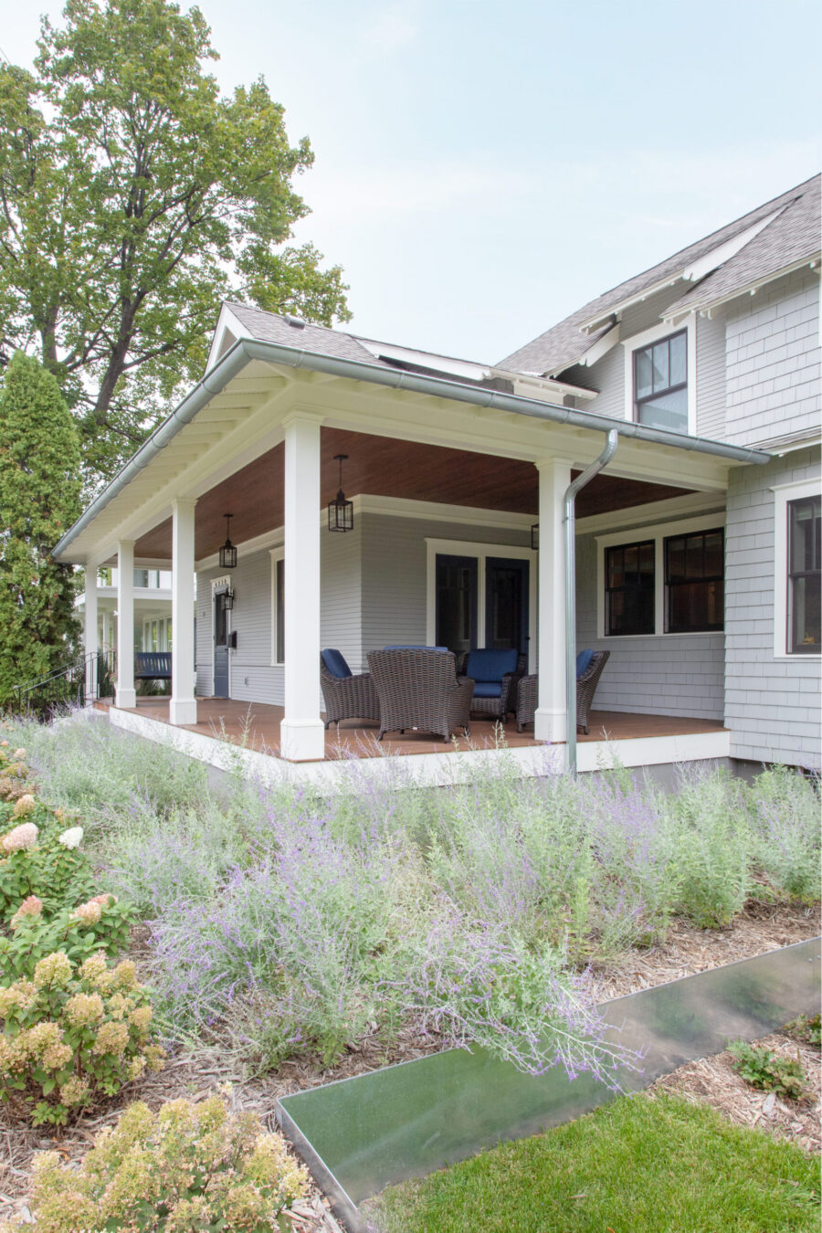 Home renovation adds gray shingled exterior, covered porch, wicker seating, beautiful gardens.