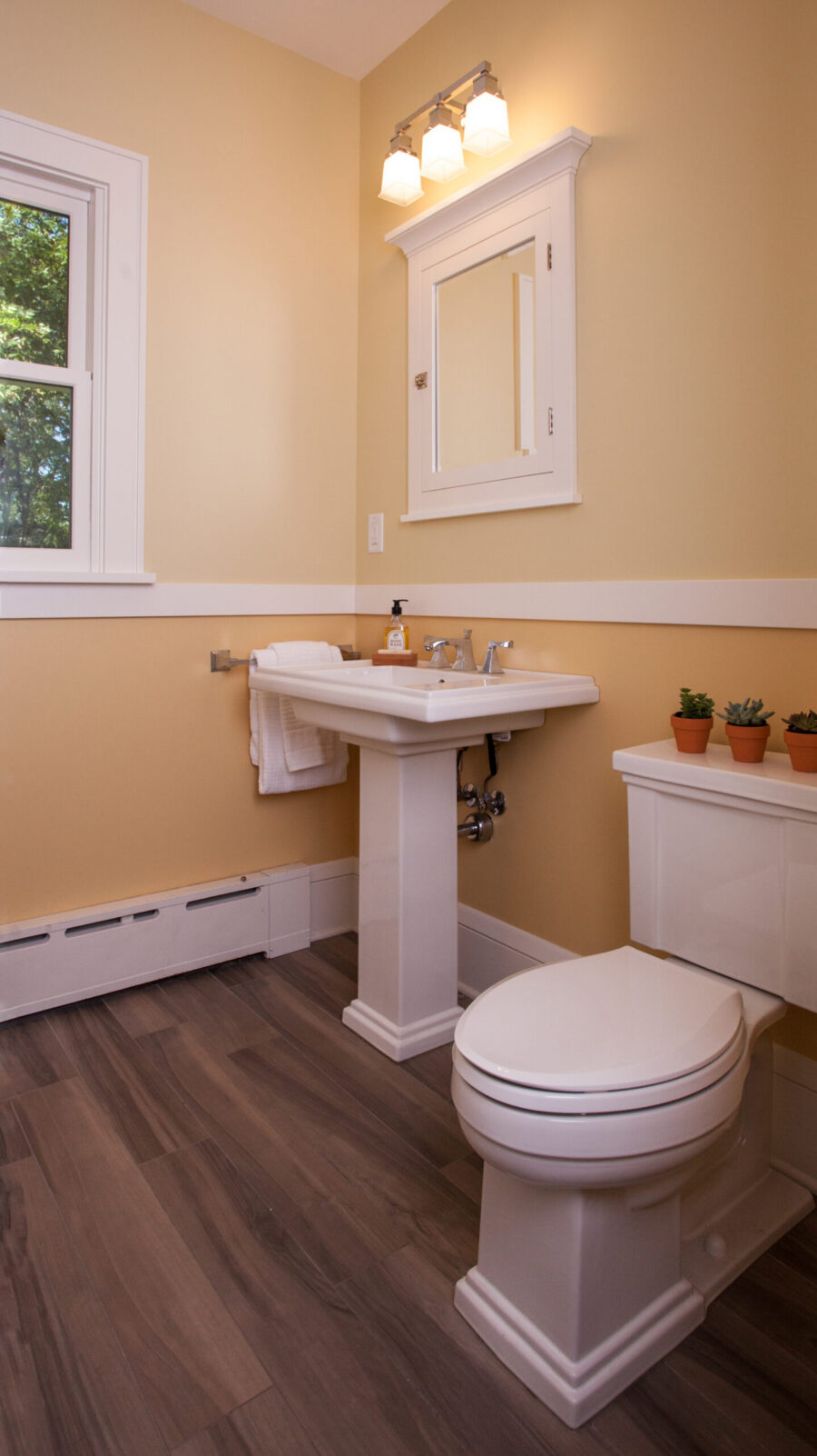 Bright Twin Cities bathroom renovation features yellow walls, white pedestal sink, toilet, and wood-look flooring.
