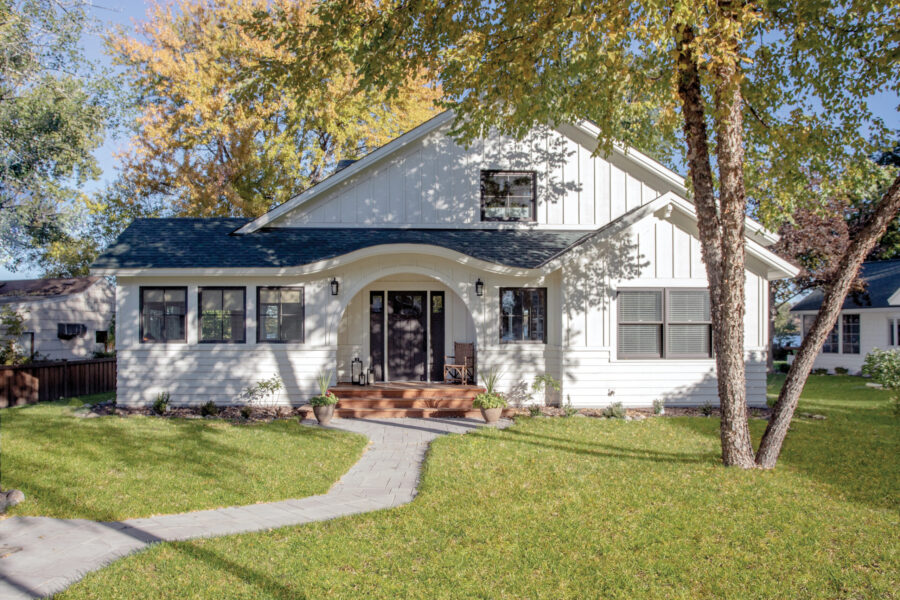 Renovated white farmhouse exterior displays dark windows, arched entry, green roof, paver path.