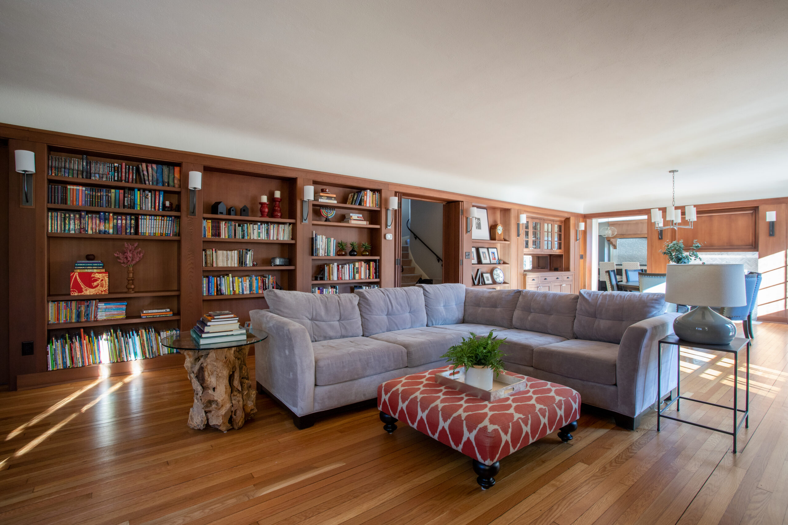 Expansive custom-built wooden bookshelves integrate sunlit hardwood floors, plush gray sectional.