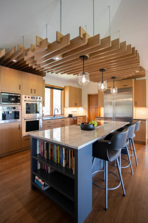 Twin Cities kitchen remodel. Slatted wood ceiling, light wood cabinets, dark floor, marble island, seating, bookshelf.