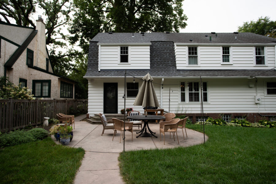 Exterior renovation yields white siding, dark gambrel roof, expansive concrete patio.