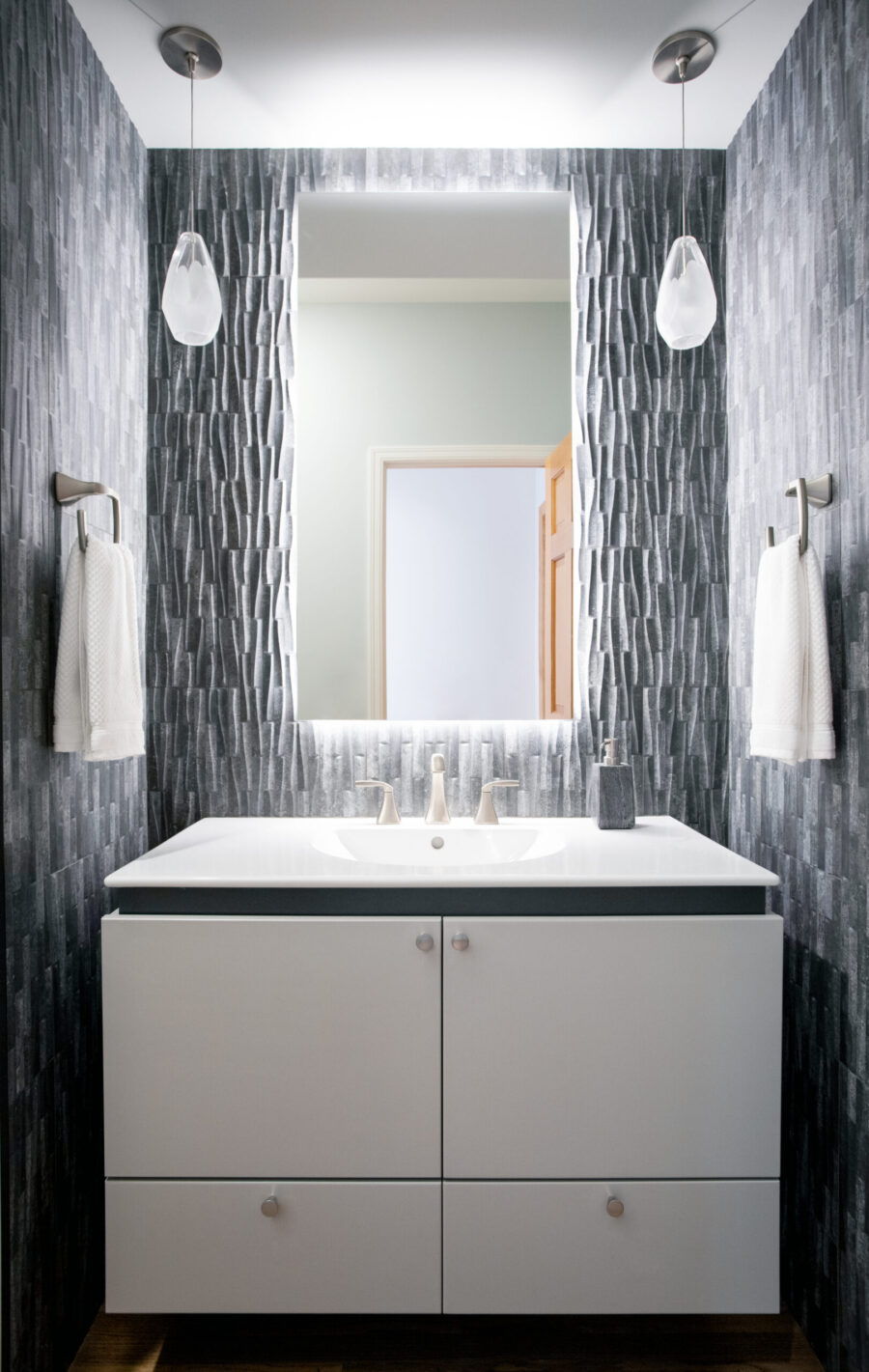 Bathroom remodel features white floating vanity, illuminated mirror, and textured gray wall tiles.