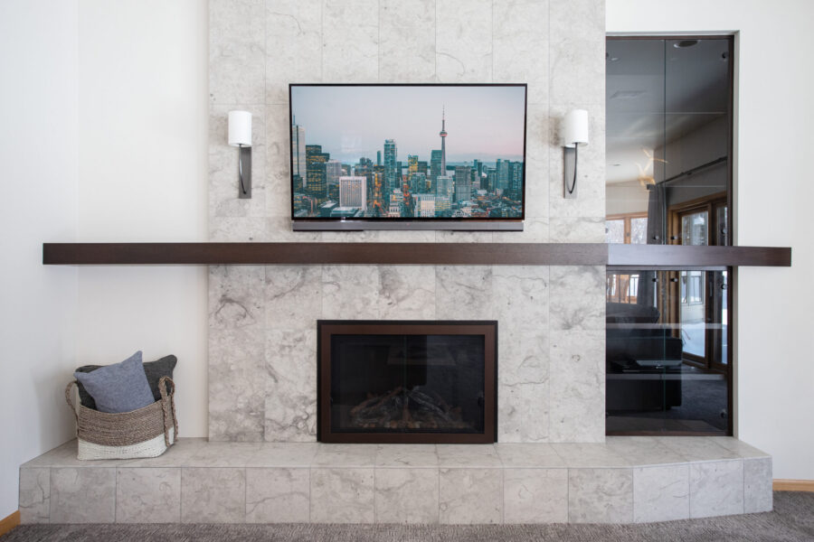 Renovation highlights grey-tiled fireplace, dark wood mantel, TV, and sconces.