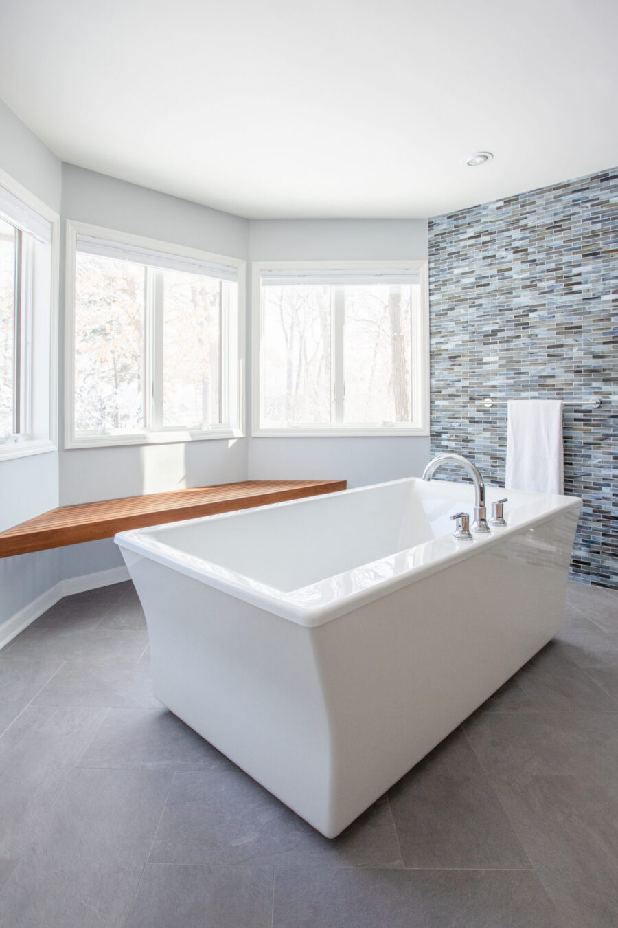 Bright Twin Cities home renovation features a spacious bathroom with a white freestanding tub, wood bench, mosaic wall.
