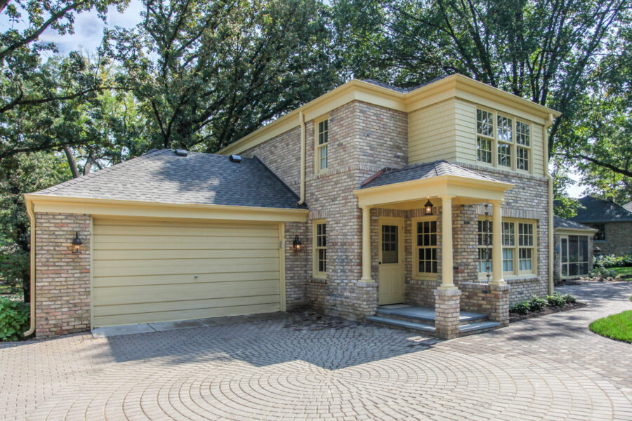 Remodeled residential exterior combines light brickwork, yellow paneling, a sheltered entryway, and patterned pavers.