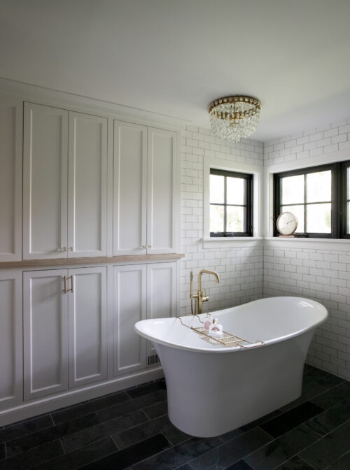 Bathroom features freestanding-tub, brass-faucet, subway-tile, chandelier, dark-floor, white-cabinets, bathroom-remodel