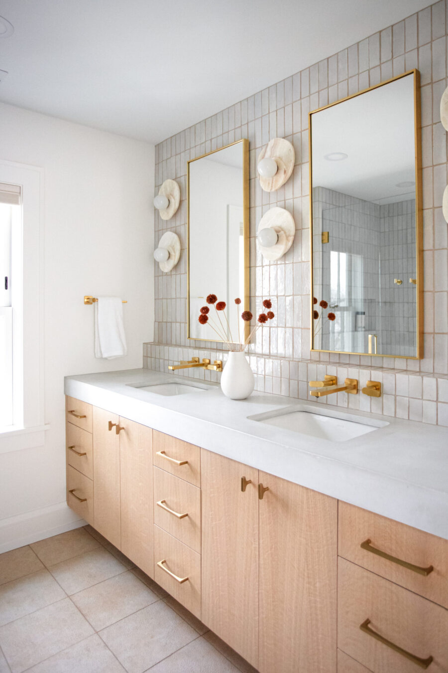 Bathroom renovation features light-wood cabinetry, white-tiled walls, gold fixtures.