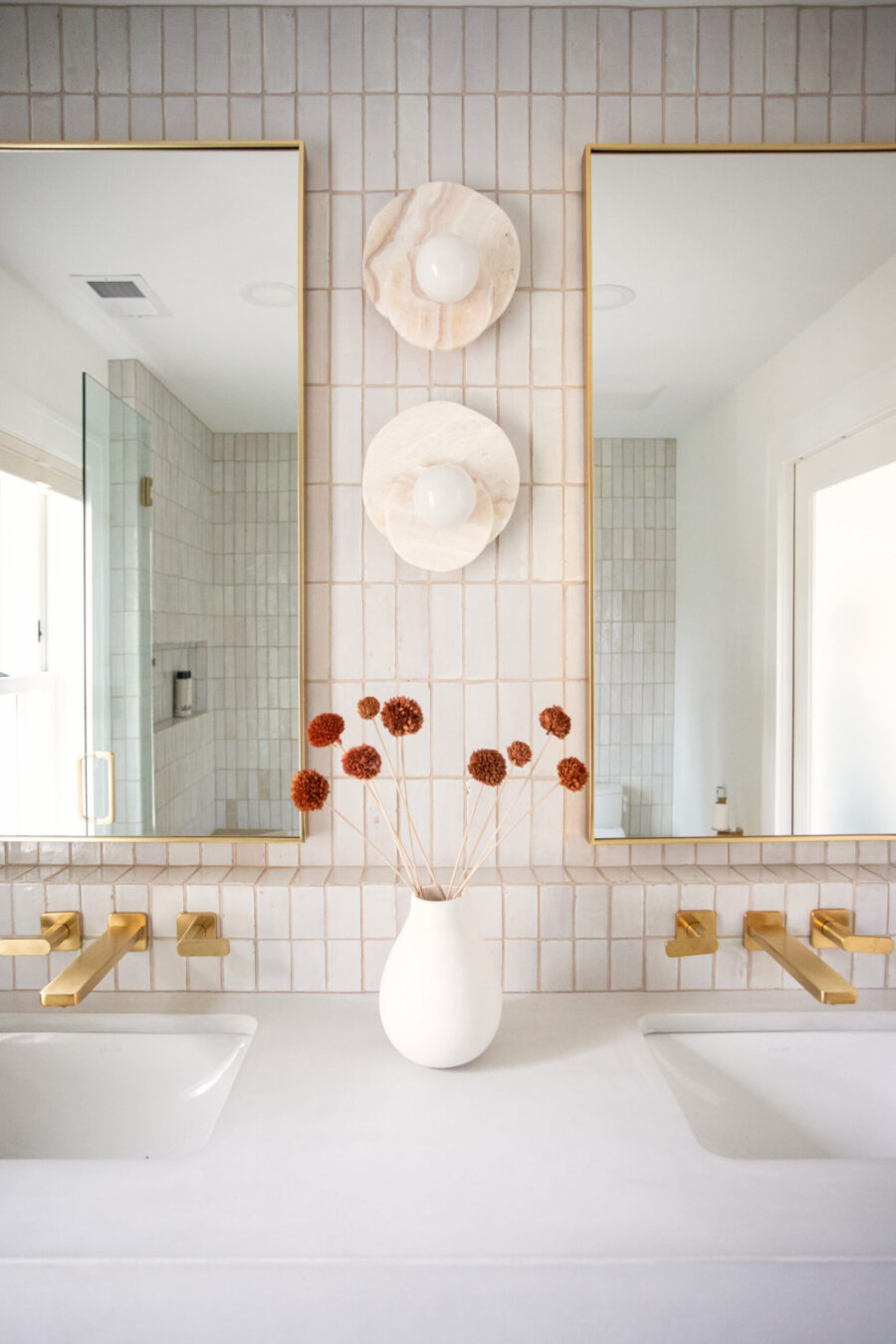 Renovated Twin Cities bathroom displays white vertical tiles, dual sinks, brass wall faucets, and round marble sconces.