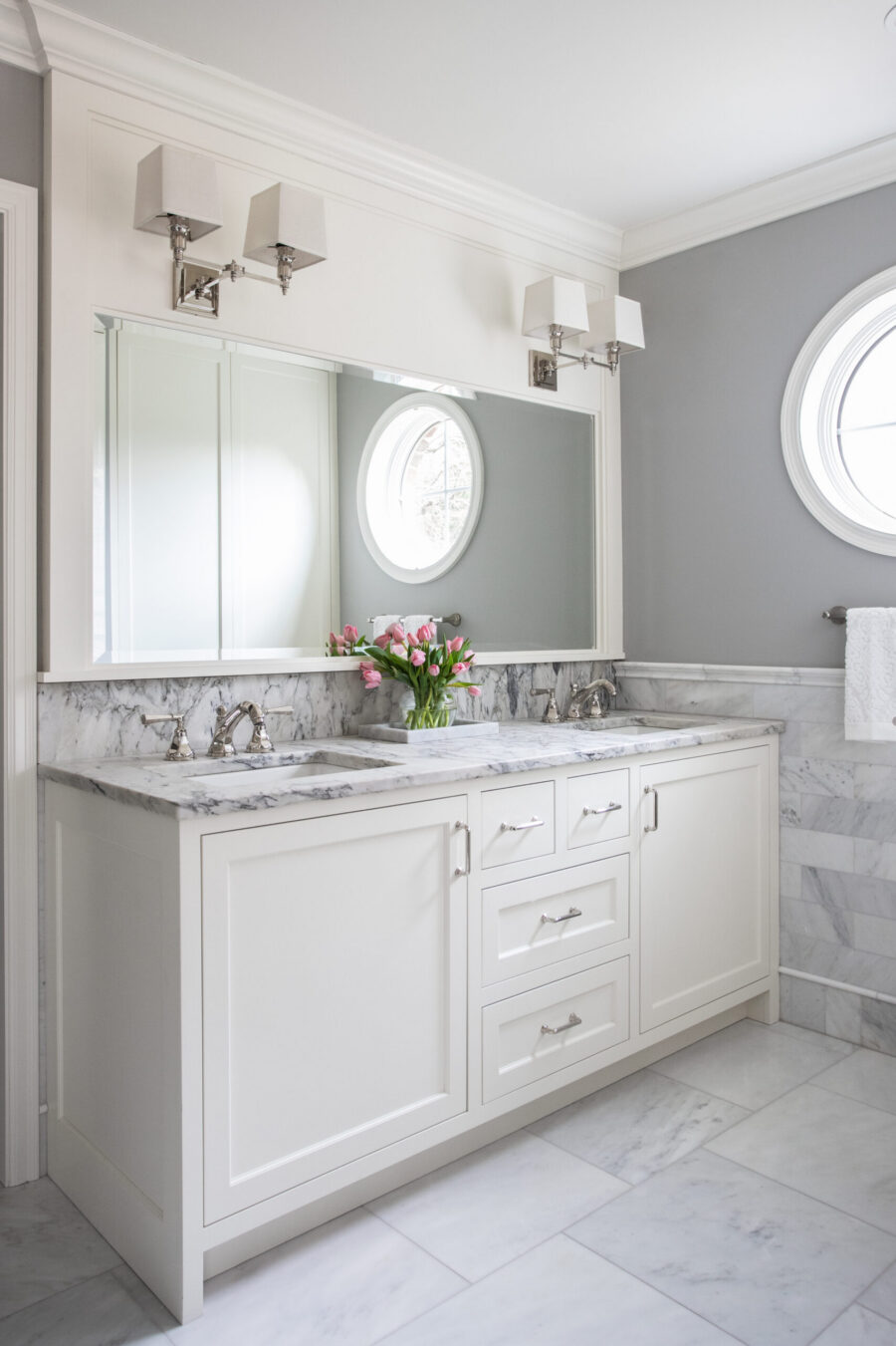 Twin Cities bathroom remodel: white marble double vanity, shaker cabinets, gray walls, bright oval windows, and sconce lighting.