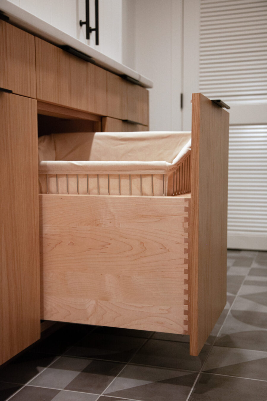 Modern light wood cabinetry features an open pull-out hamper drawer, revealing fine dovetail joinery. Twin Cities home renovation.
