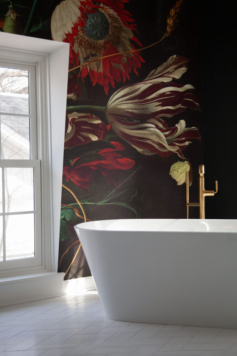White freestanding tub, dark floral wallpaper, gold faucet, bright window light.