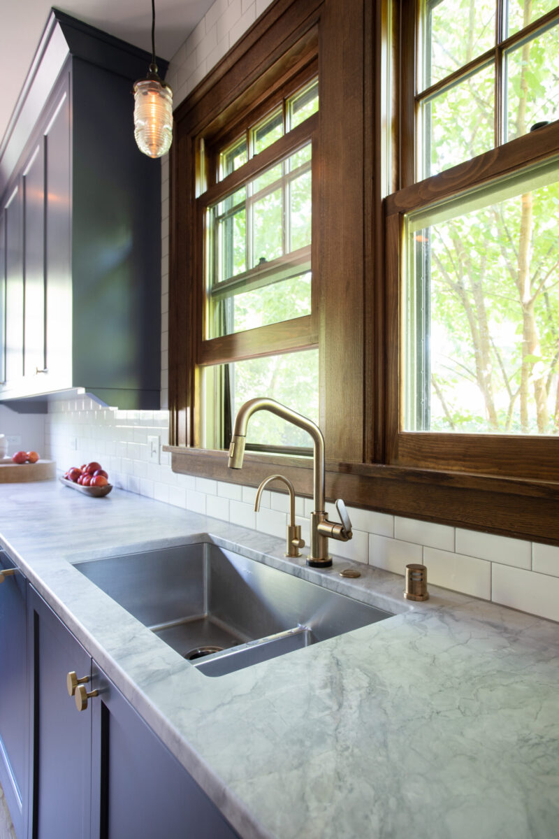 Kitchen renovation in Twin Cities features dark cabinets, marble counters, brass fixtures, and sunlit wood windows.