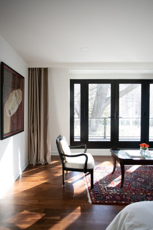 Sunlight illuminates walnut flooring, upholstered armchair, Persian rug, black-French-doors
