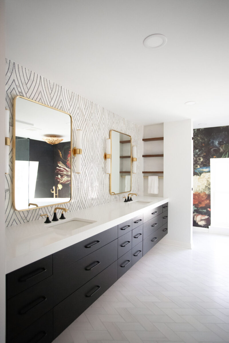 Spacious modern bathroom renovation features dark vanities, gold faucets, striking patterned wall.
