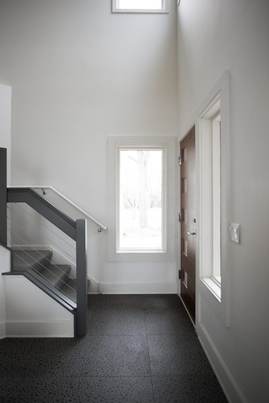 Modern Twin Cities entry hall renovation features dark patterned floor, white walls, wood door, and sleek staircase.