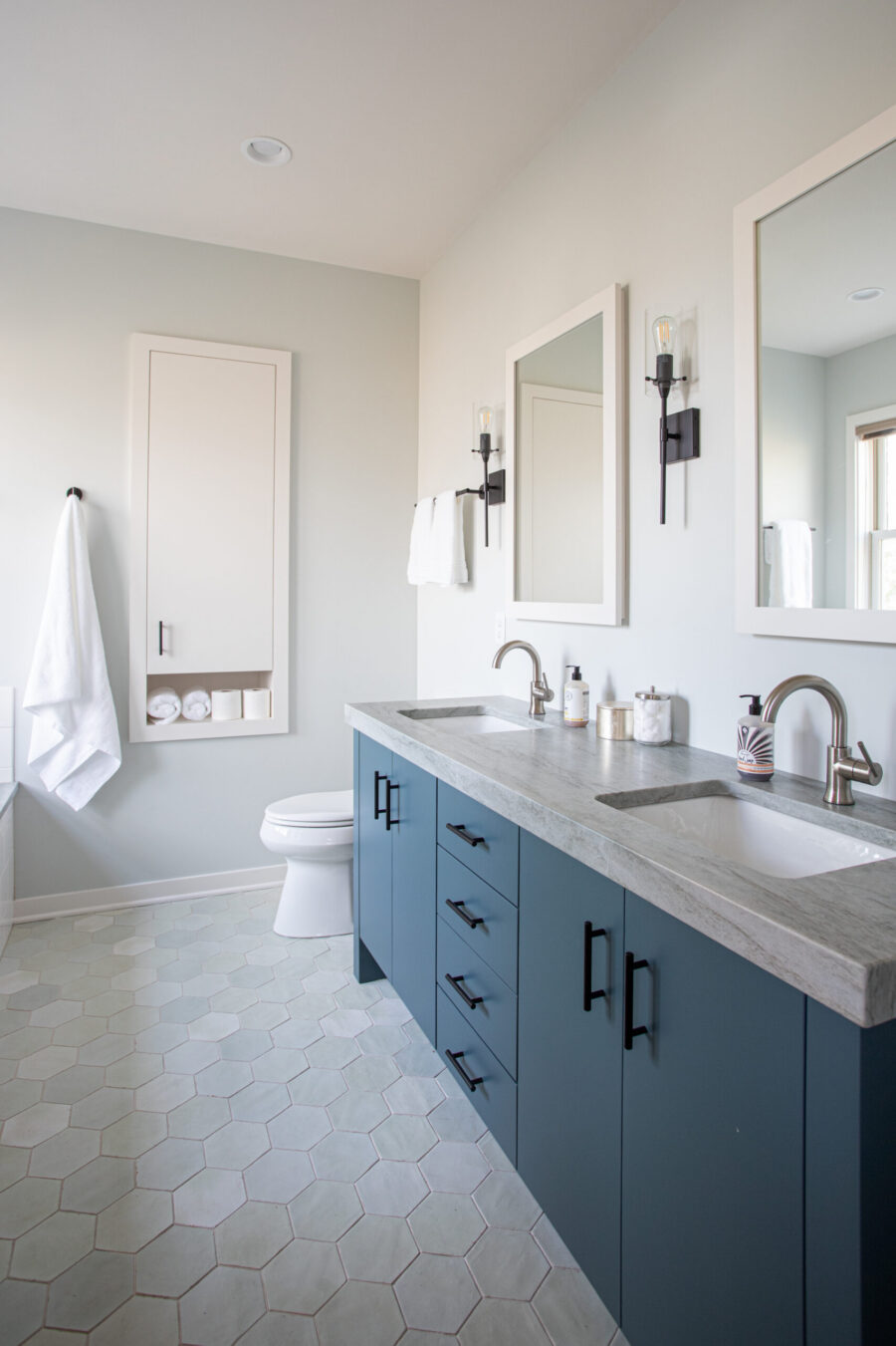 Remodeled modern bathroom has blue dual-vanity, grey stone-look countertop, hexagonal flooring.