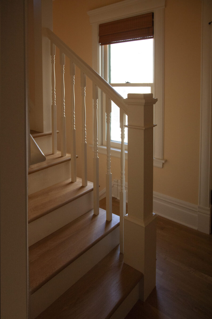 Hardwood stairs, white balusters, and a newel post reflect natural light from a blind-covered window. This home renovation enhances a Twin Cities interior.