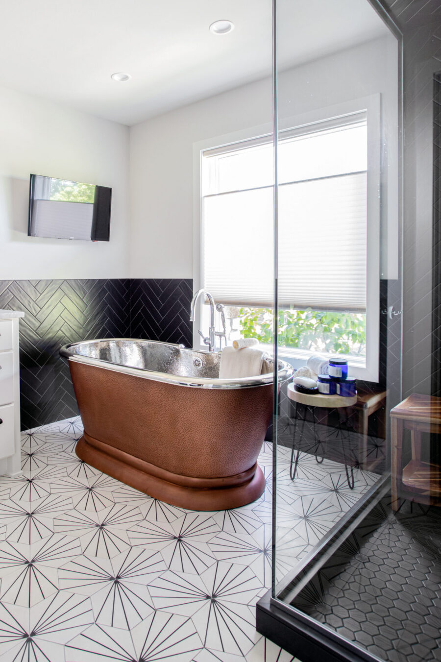 Bathroom remodel highlights hammered-copper tub, black-herringbone tiling, geometric-patterned flooring design.