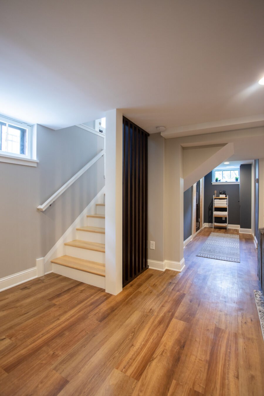 Professionally-designed basement-remodel features warm-wood-flooring, bright-white-stairs, dark-slatted-screen, gray-walls.